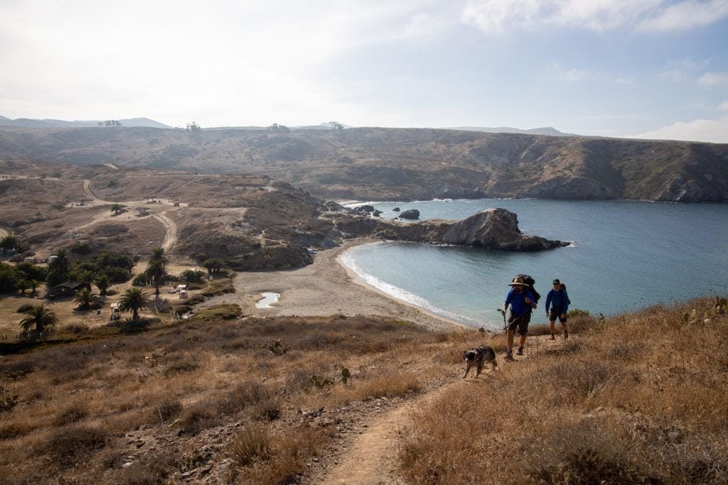 two backpackers hiking up trail on Trans-Catalina route on Catalina Island with ocean and harbor behind them