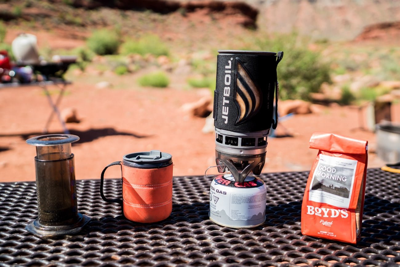 An Aeropress Go with an orange insulated mug and Jetboil Flash and orange bag of coffee grounds sitting on a campground table