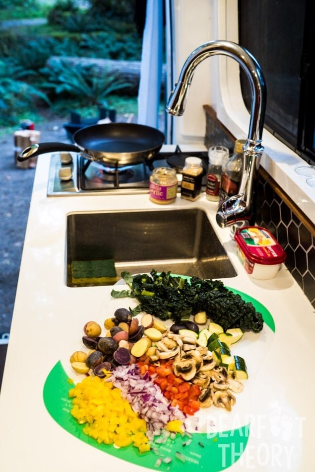 The Kitchen in Alaina's first 4x4 Sprinter camper van showing a dual propane burner, a stainless steel sink and fawcett, and a cutting board with veggies and spices