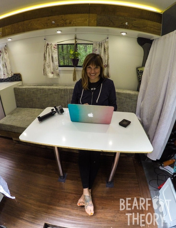 Photo of Alaina working on her laptop on the table inside her first Sprinter camper van seated on the sofa that converts to a bed