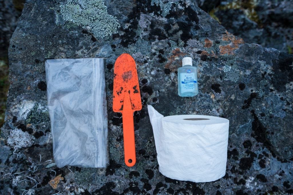 An orange trowel, toilet paper roll, hand sanitizer, and Ziploc bag laid out on the ground. This is a poop kit for backpackers.