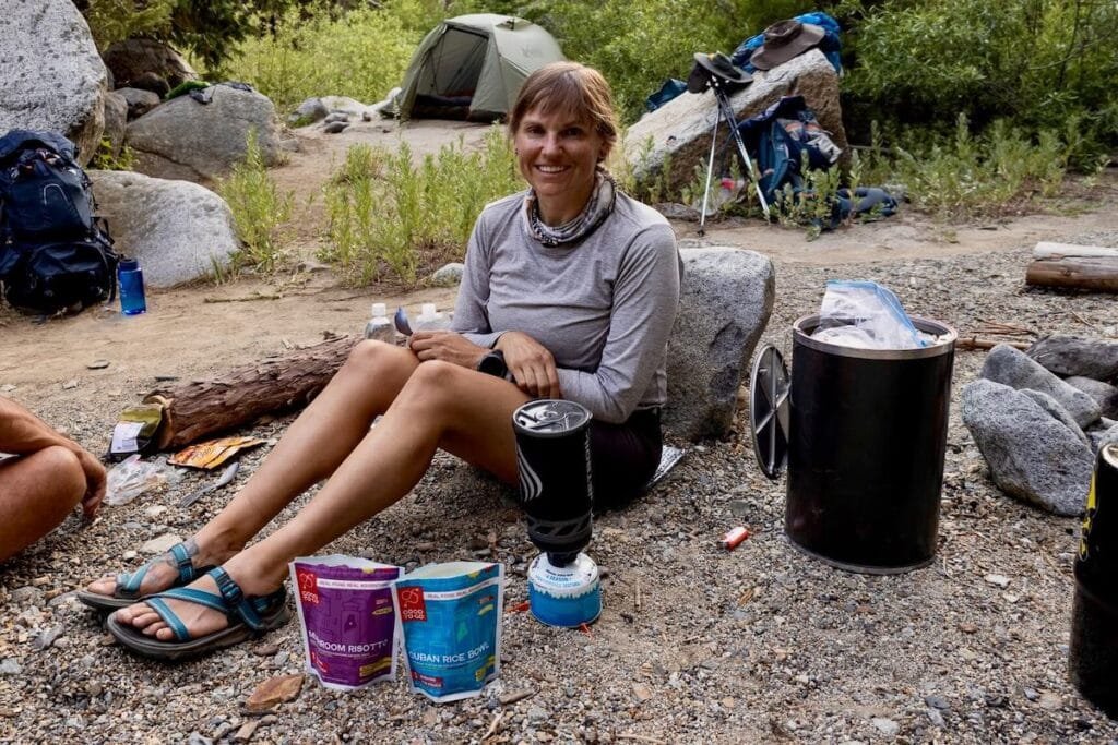 Alaina sitting on ground next to backpacking stove and bear canister on backpacking trip