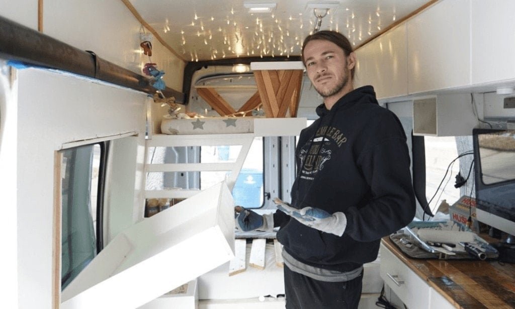 A man stands in his campervan with a fold out kids bed coming down from the wall