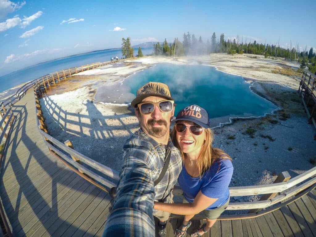 Alaina Hermiston and her partner taking a selfie next to a blue geyser in Yellowstone's West Thumb Geyser Basin with Yellowstone Lake in the Background