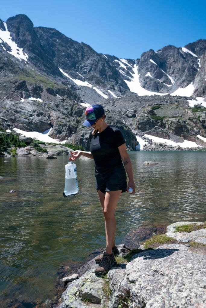Alaina walking on rock slab a lake water edge carrying water filter filled with water with mountains covered with patches of snow in background
