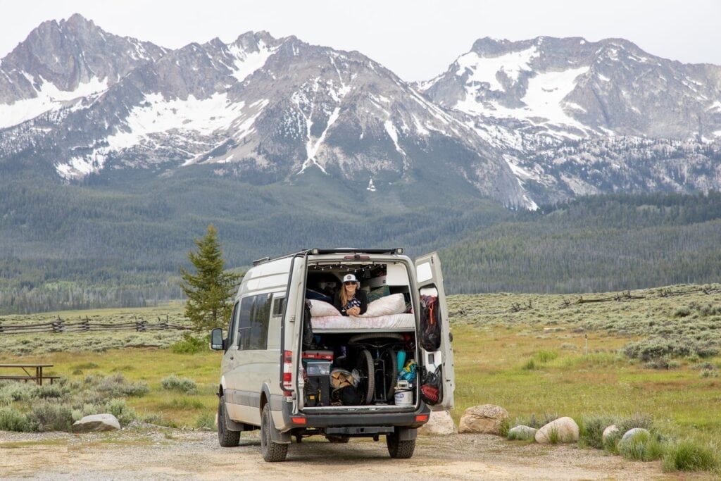 Alaina in her Sprinter van looking out the back in front of distant snowy mountains in Idaho