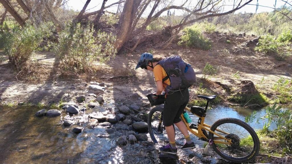 Female mountain biker with large backpack on back pushing bike across narrow and shallow river on overnight bikepacking trip