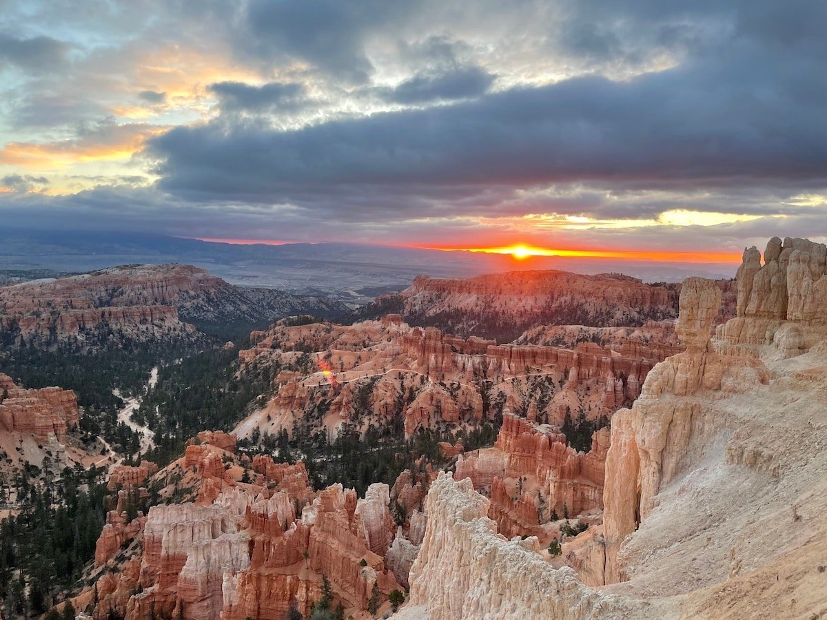 Sunrise at Inspiration Point in Bryce Canyon National Park. There is a orange and pink sun in a cloudy skies with hoodoos below.
