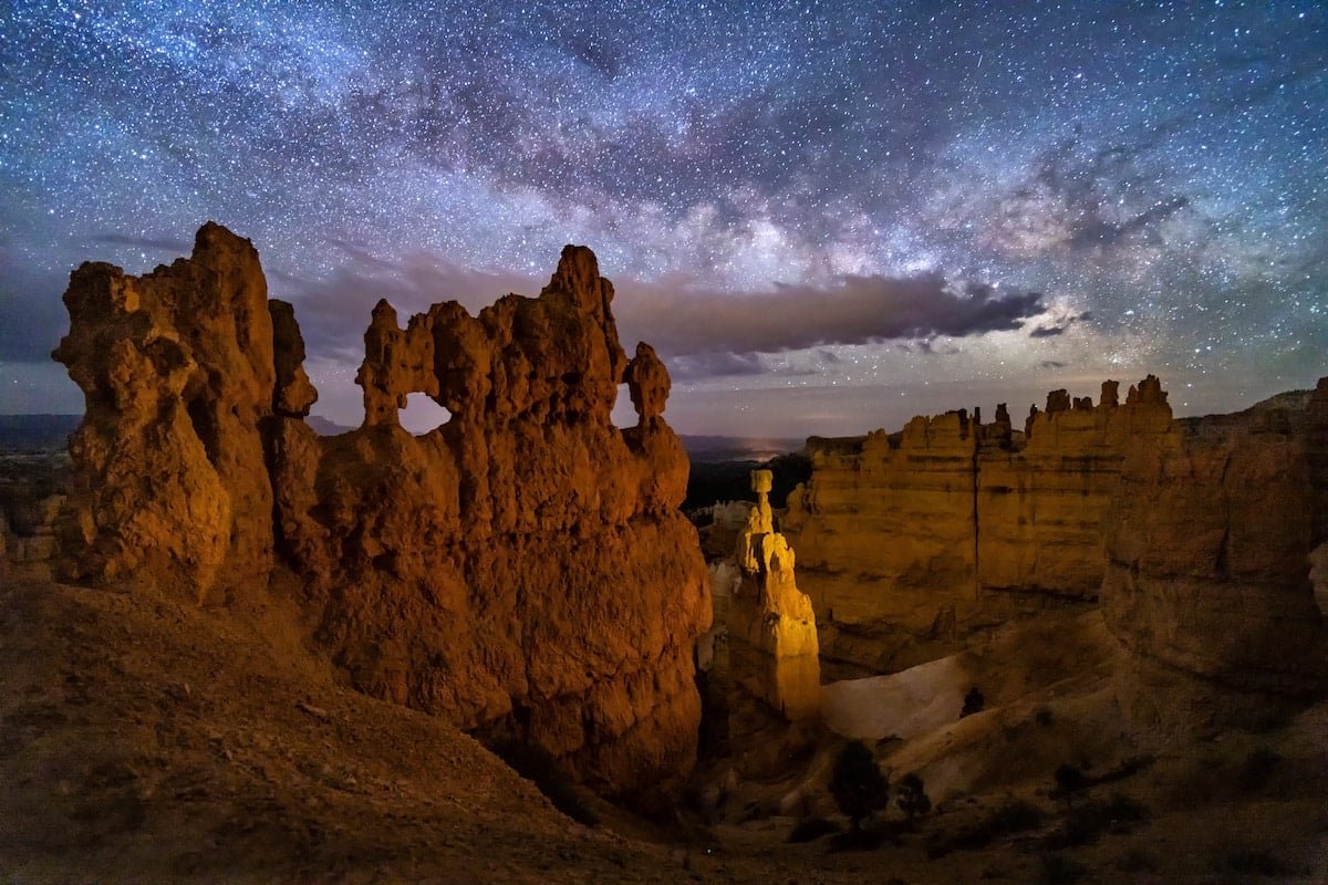 The Milky Way with stars and clouds over an orange rock formation at Bryce Canyon National Park at night.
