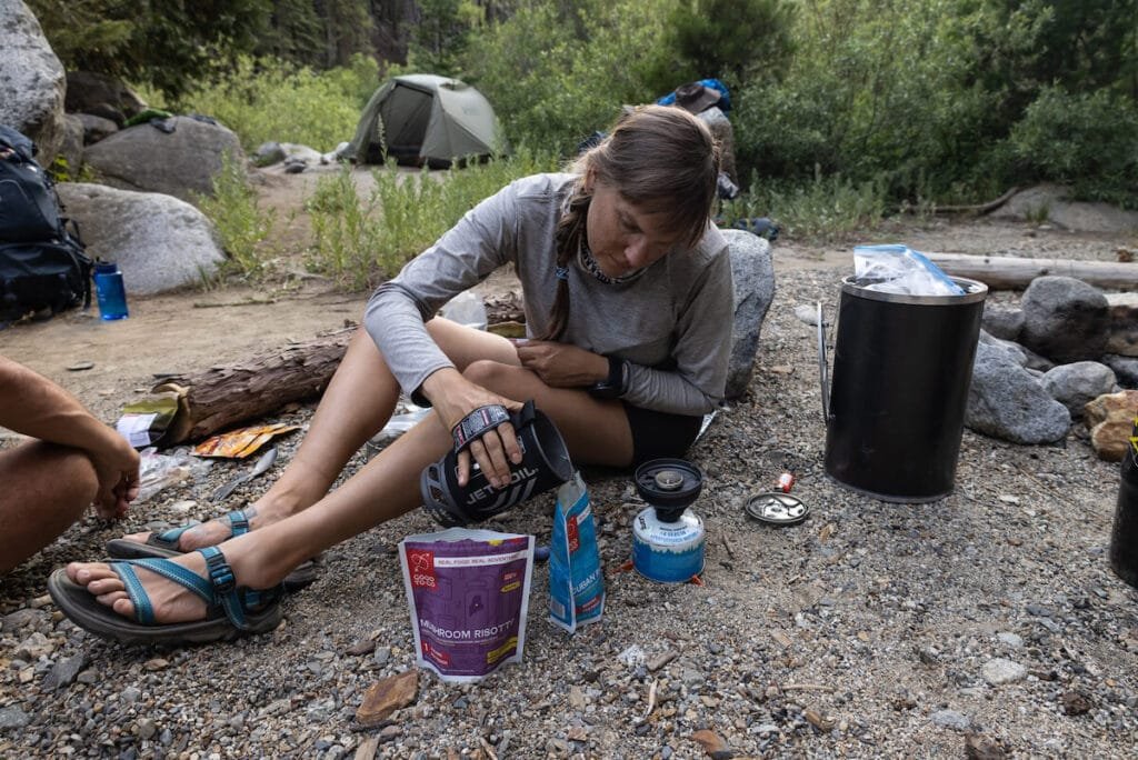 Alaina pouring hot water from Jet Boil backpacking stove into dehydrated meal pouch on a backpacking trip with backpacking gear spread out around her
