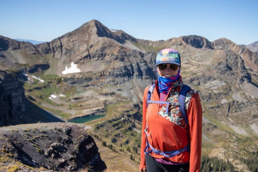 Alaina smiling for photo on hike Colorado wearing hat, buff, sunglasses, and long sleeve shirt