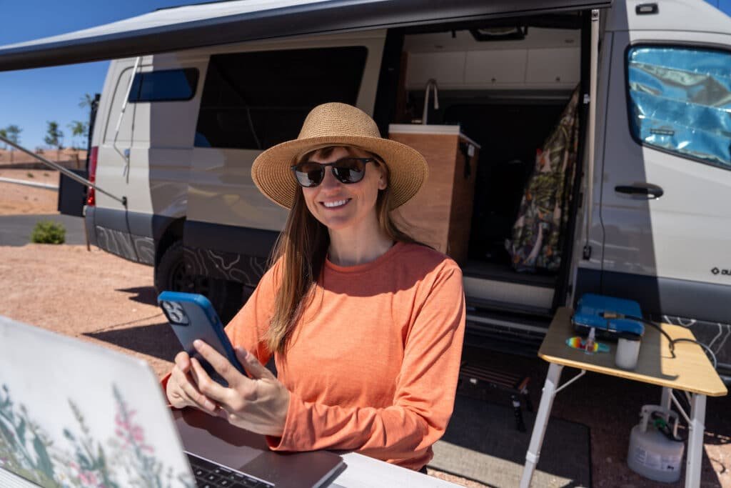 Alaina Hermiston with a laptop and her phone in her hand sitting at a picnic table that is next to a sprinter van