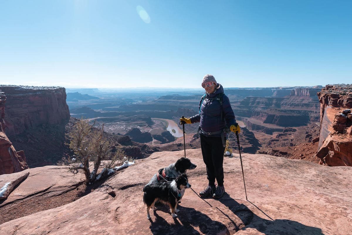 Alaina Hermiston with hiking poles and dogs standing at Dead Horse State Park lookout over Colorado River and Canyonlands National Park in Utah