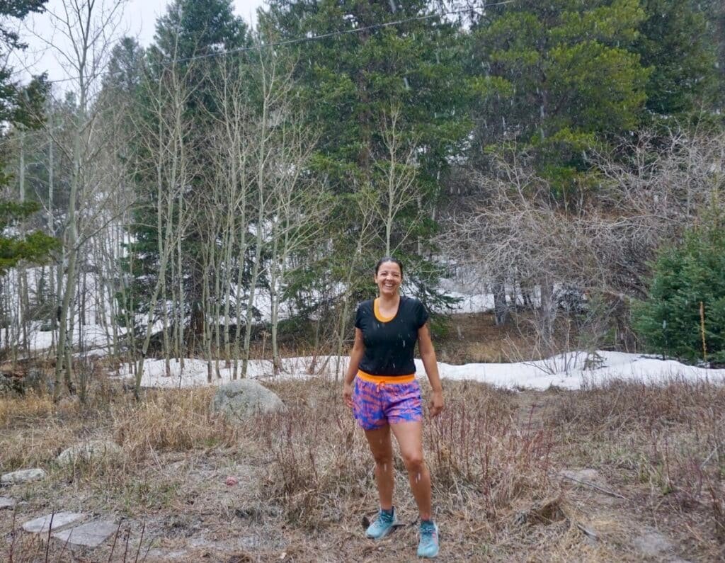 A woman smiles at the camera wearing Janji AFO 3" Shorts. There's a partially snowy trail in the background