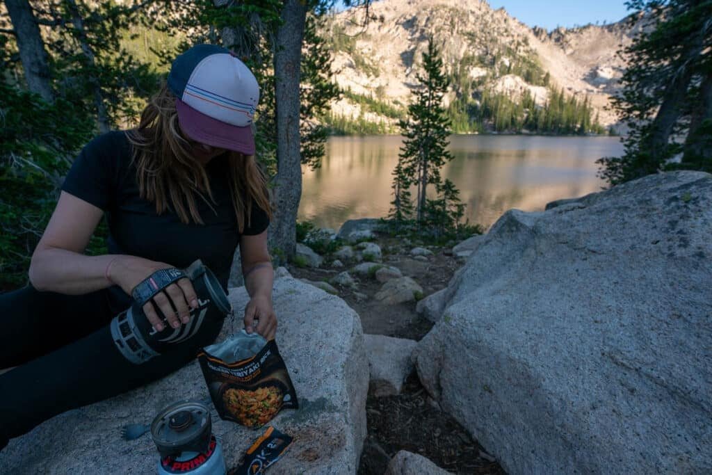Alaina Hermiston pouring water from a Jetboil into a backpacking meal pouch next to a mountain lake