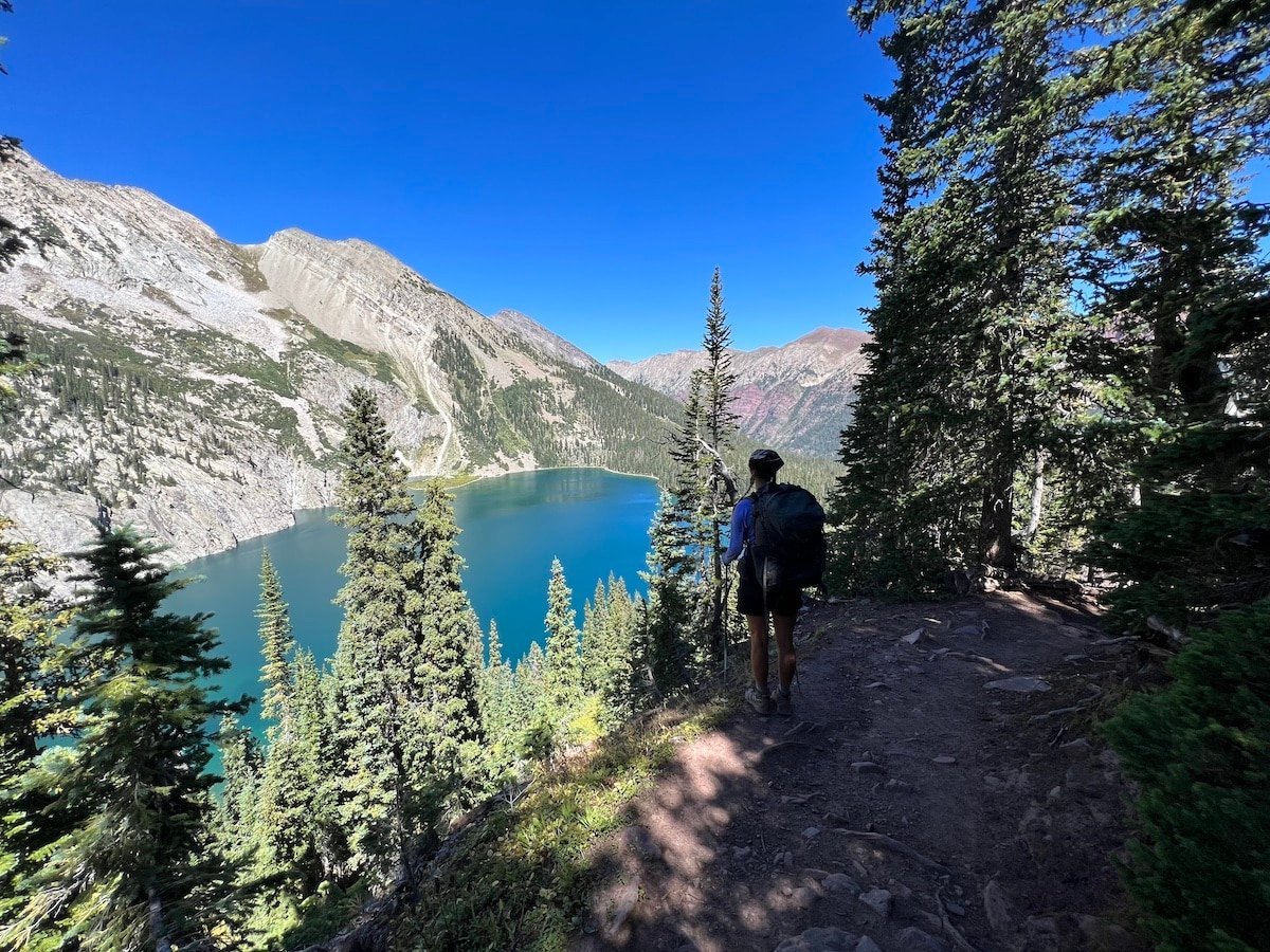 Hiker looking down on Snowmass Lake surrounded by mountains and trees in the forest.