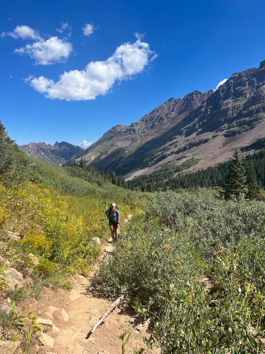 Female hiker with greenery along the trail and mountain peaks in the distance behind her