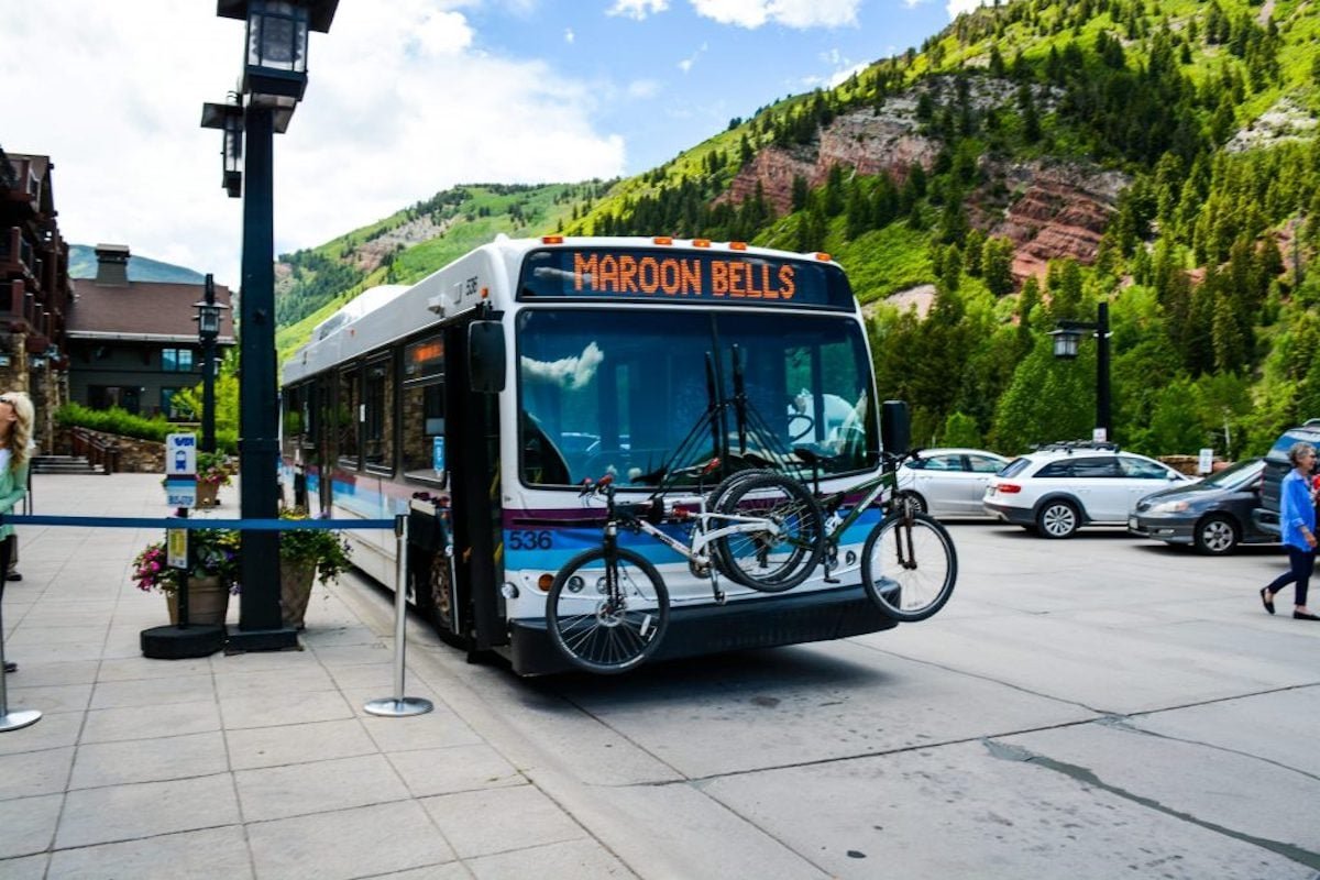 Shuttle that says Maroon Bells on it with bikes attached to the front and a green hill in the background