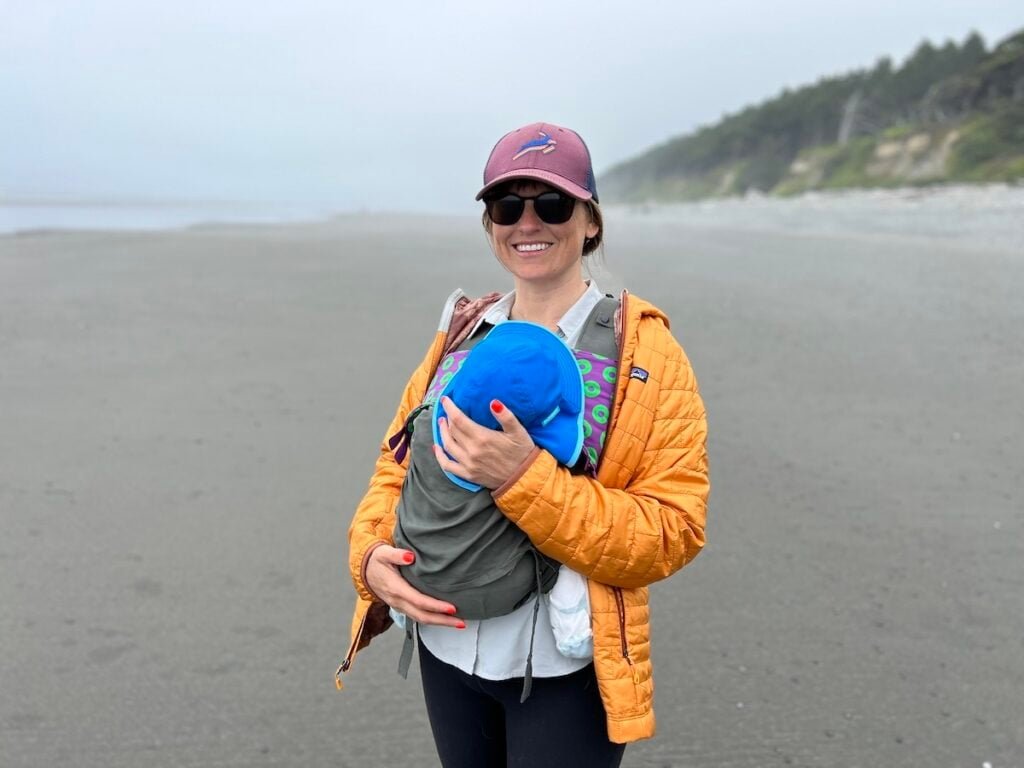 Terrain Spark founder Alaina Hermiston on a foggy beach in Olympic National Park wearing a gold Patagonia Nano puff and holding her baby in a carrier