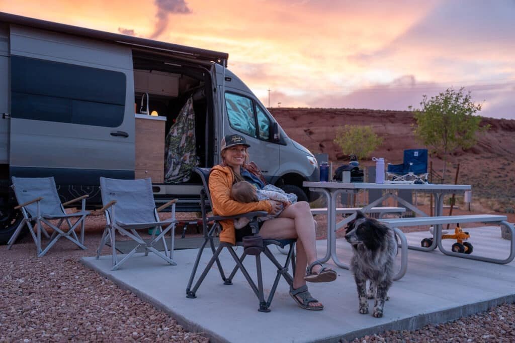 Alaina Hermiston sitting in a camp chair holding baby at a campsite at Roam Horseshoe Bend RV Resort next to a Sprinter Van