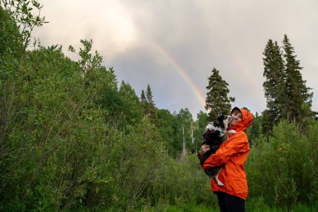 Alaina Hermiston wearing a red Arc'teryx Beta SL Rain Jacket holding her dog standing underneath a rainbow